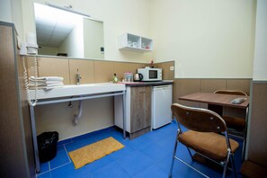 Una kitchenette compacta con lavamanos, microondas y un refrigerador pequeño. Se observa una silla y una mesa con una silla, además de un bote de basura.