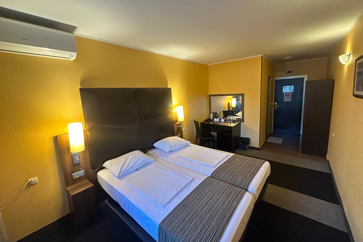 A hotel room with a bed, bedside lamps, a desk with a chair, a mirror, and a door.