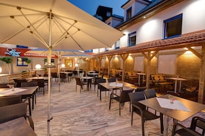 An outdoor dining area with tables and chairs, covered by umbrellas, and a building with signage in the background. - Hotel Leimer Brv U (Lenzing)