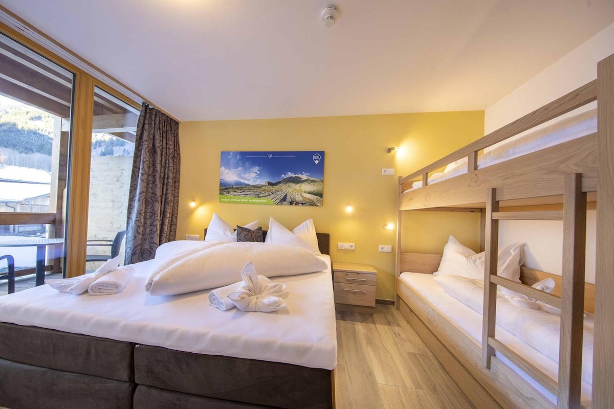 Una habitación de hotel con cama, mesita de noche, litera de madera, ventana con cortinas y un cuadro en la pared.