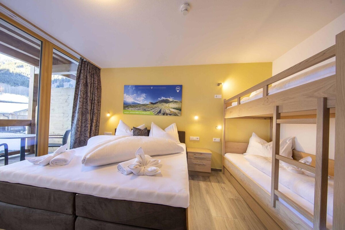 A hotel room with a bed, a nightstand, a wooden bunk bed, a window with curtains, and a painting on the wall.