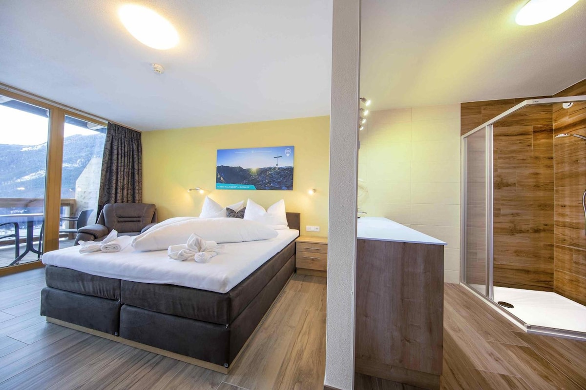 A modern hotel room with a large bed, a seating area, a shower, and a view of the mountains.