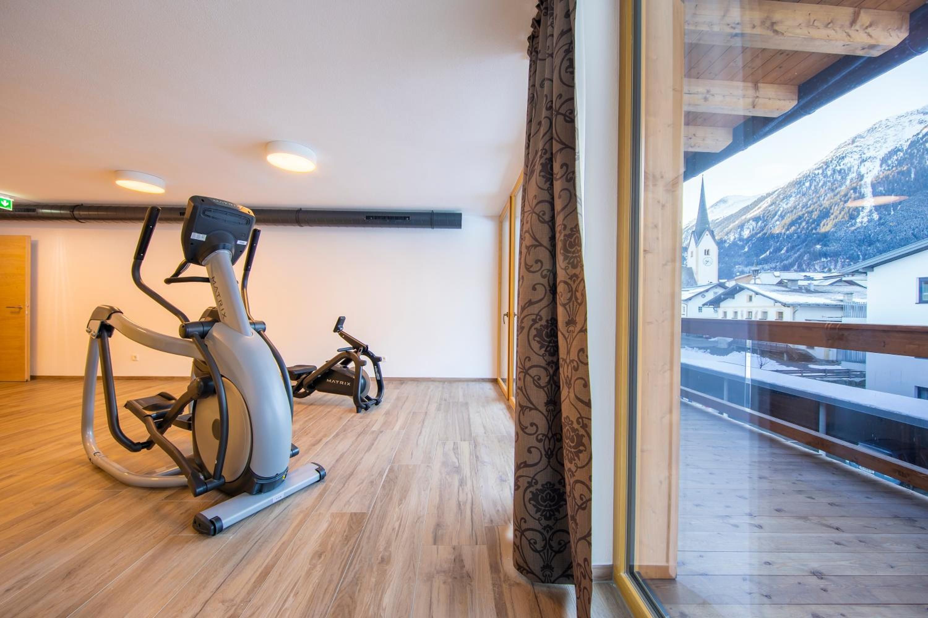 A room with a treadmill and stationary bike, wooden flooring, a large window with a view of a snowy mountain, and a wooden ceiling.