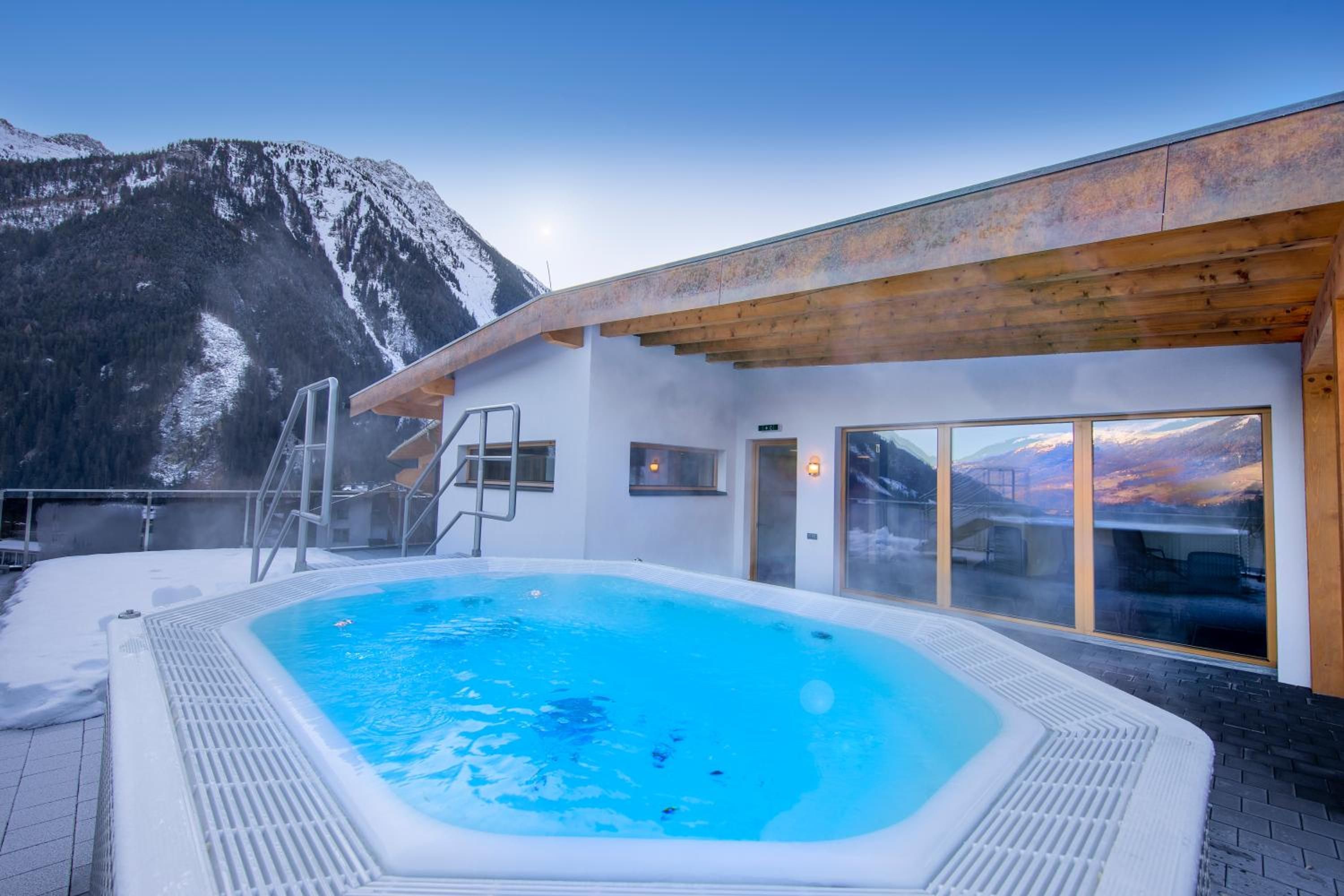 Een hottub met uitzicht op de bergen, een houten terras en een glazen deur naar een kamer.
