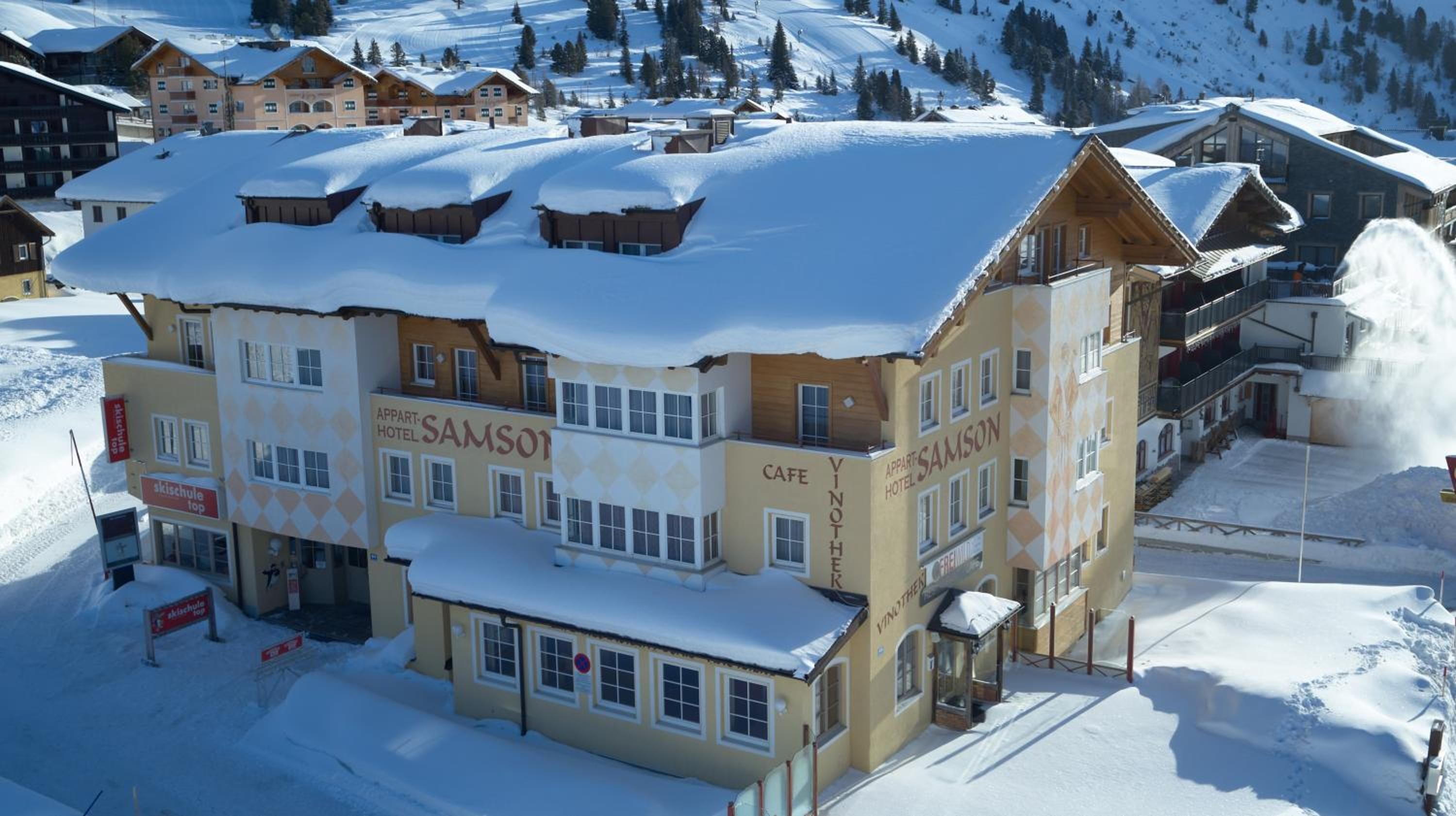 A multi-story building with 'Appart Hotel Samson' signage, a cafe, and ski chalets in a snowy mountainous area.