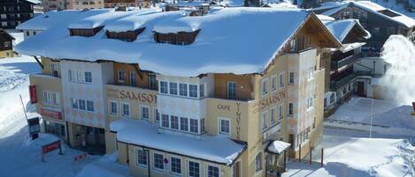 Un edificio de varios pisos con la señalización 'Appart Hotel Samson', una cafetería y chalés de esquí en una zona montañosa nevada.