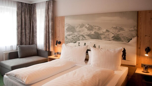 Habitación de hotel con cama, sofá, un cuadro de montañas y ventana con cortinas.