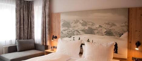 Habitación de hotel con cama, sofá, un cuadro de montañas y ventana con cortinas.
