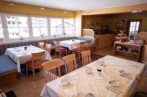 A dining area with multiple tables and benches, large windows with views, and a kitchen area visible through an open door.