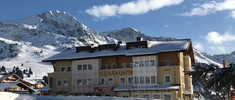A multi-story building with a sign for 'Appart Hotel Samson' and 'Cafe Yvonnthe' in a snowy mountainous area.