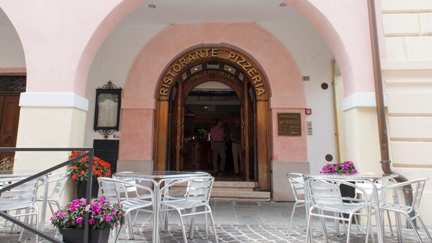 L’entrée d’une pizzeria, avec une porte en arc de cercle, des tables à l’extérieur avec des chaises en métal, et une enseigne indiquant le nom de l’établissement.