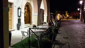 Un espace extérieur aménagé pour les repas, avec des tables et des chaises, une rue pavée et une entrée d’immeuble avec une enseigne.