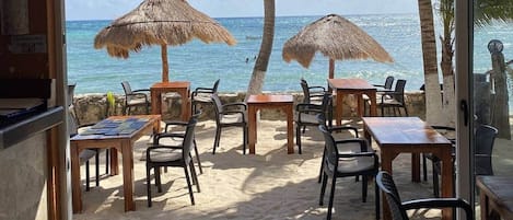 An outdoor seating area with tables and chairs, thatched umbrellas, and a view of the ocean.