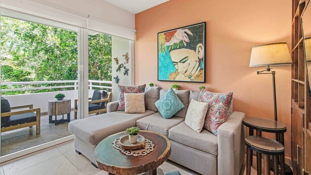 A living room with a sofa, a coffee table, and a floor lamp. There is a painting on the wall and a balcony with a view of greenery. - Akumal Bosque | 8 Min Walk To Beach | Rooftop Pool (Grand Sirenis)
