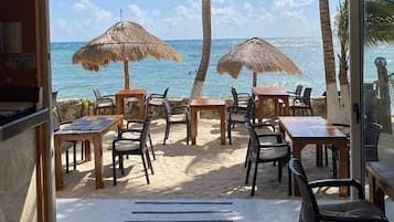 Een terras met tafels en stoelen, rieten parasols en uitzicht op de oceaan.