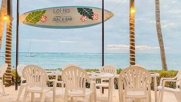 A snack bar with a view of the ocean, surrounded by white plastic chairs and tables.