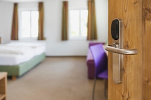 Una habitación de hotel con una puerta de madera, una cama, un sillón morado y una ventana con cortinas.