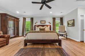 A bedroom with a large bed, a ceiling fan, a chair, and a view of greenery through the window. - Private 7-Acre Luxury Knoxville Retreat. Sleeps 14. Theater + Game Room (Knoxville)