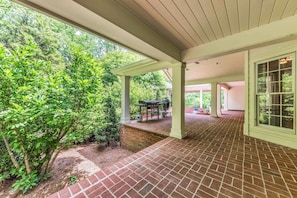A covered patio area with brick flooring, a grill, and a view of a lush garden.