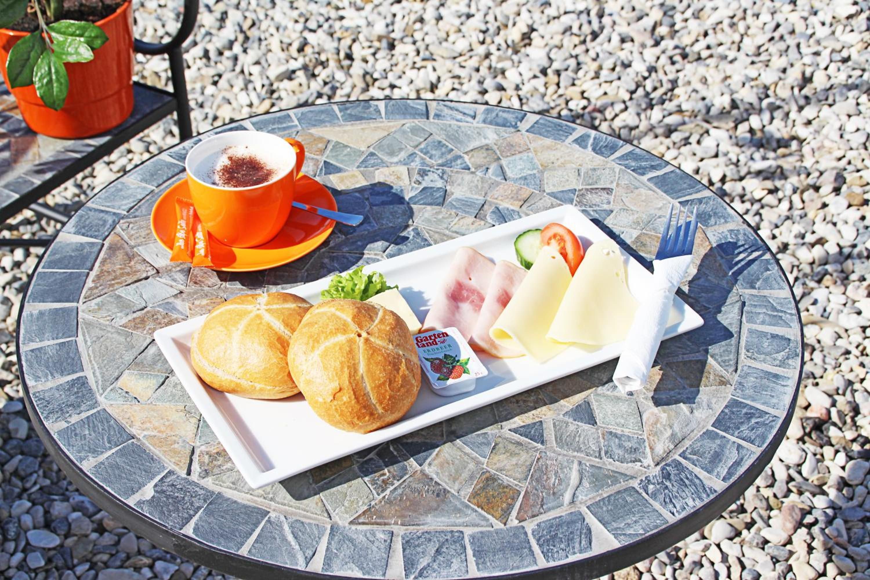 A round table with a mosaic tabletop, a plate with bread, cheese, and a small container of yogurt, a cup with a spoon, and a plant in a red pot.