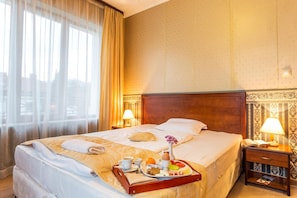 Habitación de hotel con una cama grande, una mesita de noche con lámpara, una bandeja con productos para el desayuno y una ventana con cortinas traslúcidas.