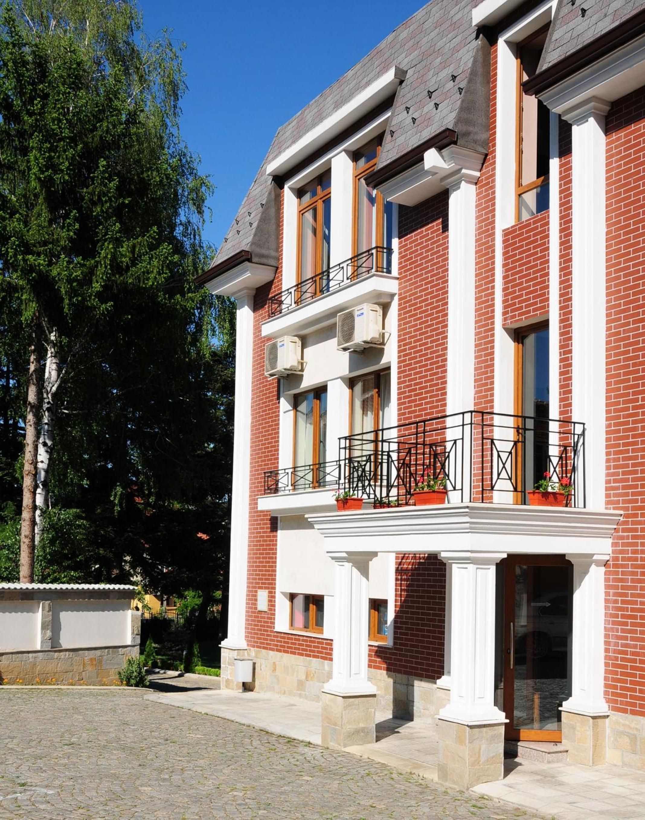 A two-story building with a brick facade, white trim, and a balcony with potted plants.