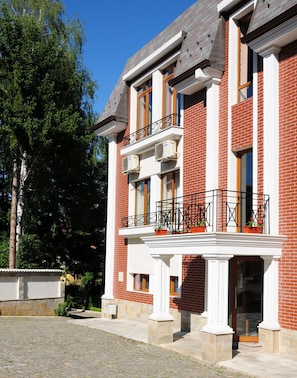 A two-story building with a brick facade, white trim, and a balcony with potted plants.