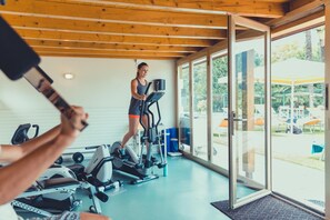 A gym with cardio machines, a person using an elliptical, and a view of the outdoors through large glass doors. - Hotel Tobler (Ascona)
