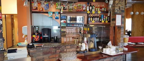 Zona de bar con mostrador de ladrillo, estantes con aperitivos y bebidas, una máquina de café y un baño visible al fondo.