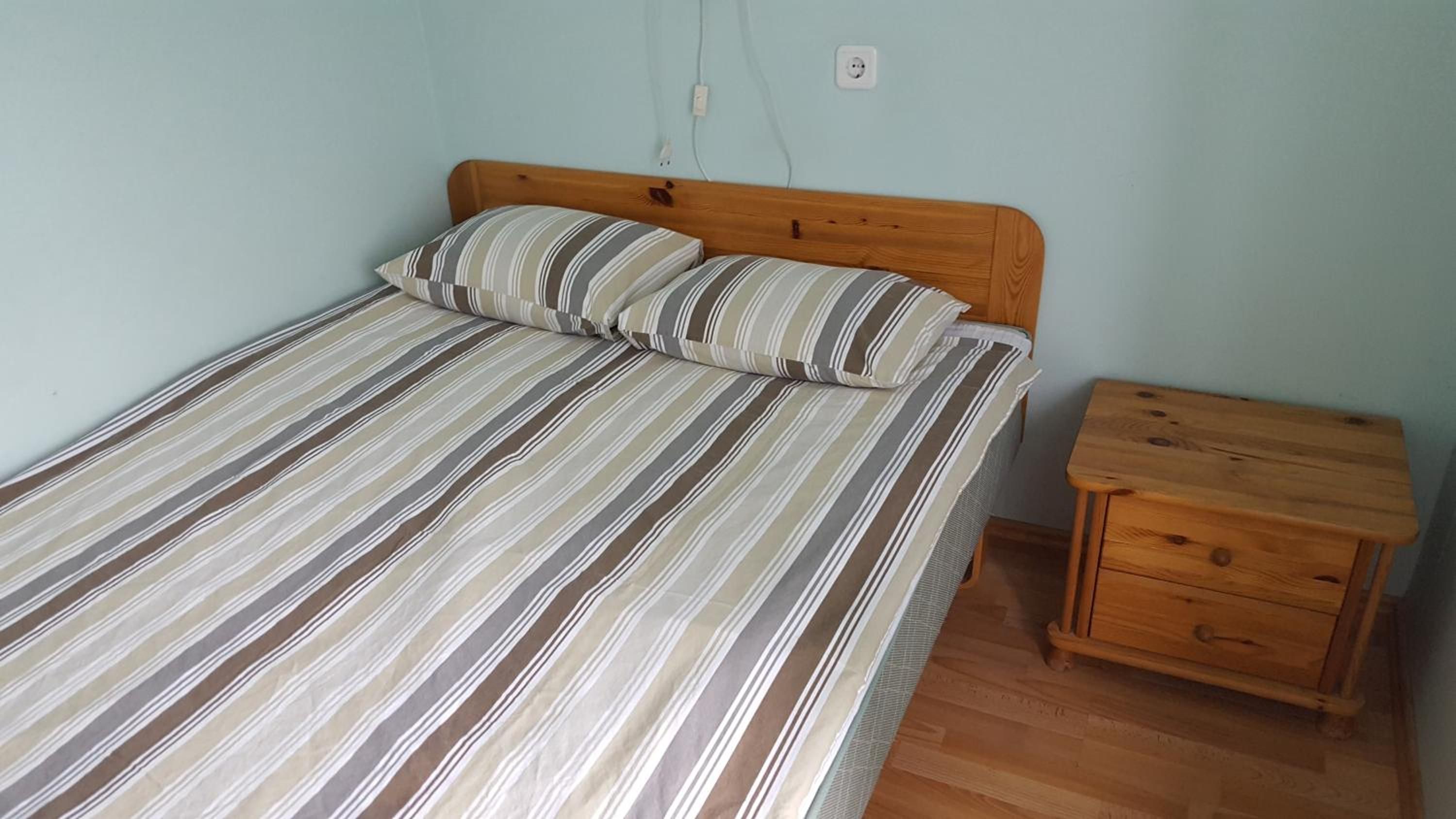 A single bed with a striped bedspread and two pillows, a wooden nightstand with a drawer, and a wall socket.