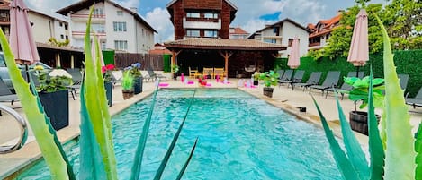 Una piscina con sillones de descanso y sombrillas, rodeada de edificios con techo de estilo alpino.