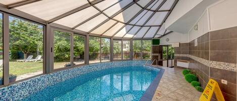 A covered swimming pool with a glass ceiling, surrounded by a tiled wall and a view of the outdoors.