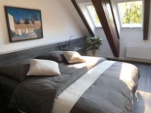 Un dormitorio con una cama grande, una mesita de noche, una planta en maceta y un cuadro en la pared.