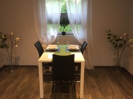 Un comedor con una mesa dispuesta para dos, una ventana con cortinas translúcidas y dos plantas en macetas a cada lado.
