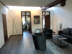 Una sala de estar con un sofá, una mesa de centro y una silla. Hay una puerta que da a una cocina y un pasillo.