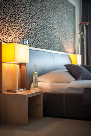 Quarto de hotel com cama, mesa de cabeceira com abajur e papel de parede estampado.