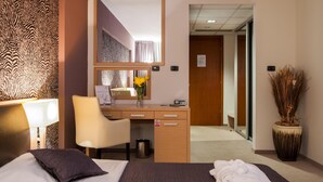 Habitación de hotel con cama, escritorio, silla, espejo, cesto de basura y planta en maceta.