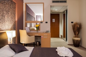 Habitación de hotel con cama, escritorio, silla, espejo, cesto de basura y planta en maceta.