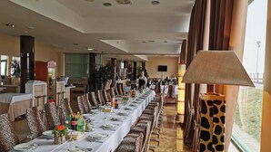 Una larga mesa de comedor con sillas de estampado de leopardo, un camarero con uniforme y un ventanal con cortinas.