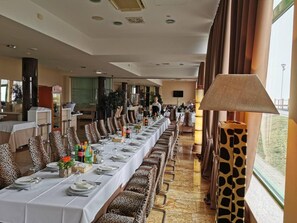 Una larga mesa de comedor con sillas de estampado de leopardo, un camarero con uniforme y un ventanal con cortinas.