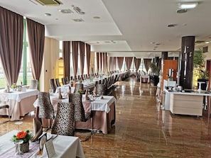 Uma longa sala de jantar com mesas e cadeiras, grandes janelas com cortinas e piso lustroso.