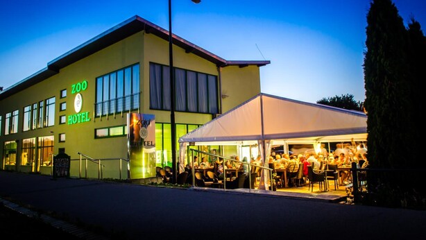Un edificio con un letrero que dice 'HOTEL', un letrero que dice 'ZOO' y un área al aire libre con carpas donde personas están cenando.