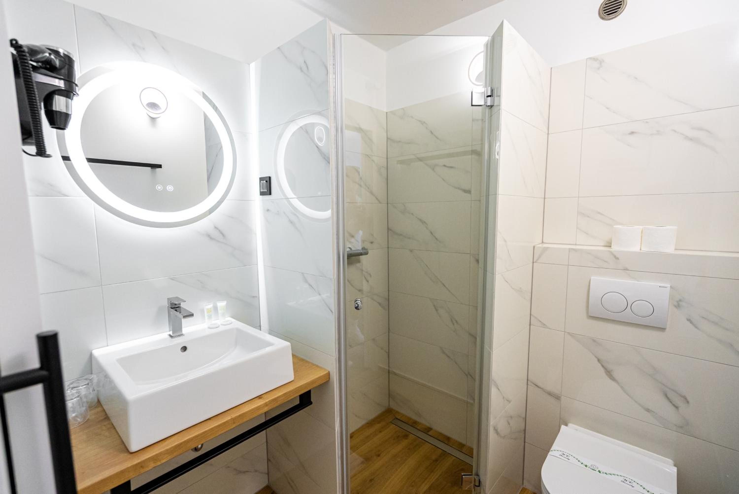 A modern bathroom with a white sink, a mirror with a circular light, a glass shower enclosure, and a toilet.