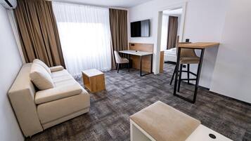 Ein modernes Hotelzimmer mit einer Couch, einem kleinen Holztisch, einem Schreibtisch mit Stuhl, einem an der Wand montierten Fernseher und Blick durch das Fenster.
