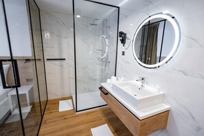 Un baño moderno con ducha envuelta en vidrio, tocador de madera con lavamanos blanco y espejo redondo con luz.