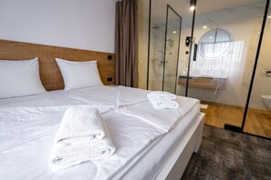 Habitación de hotel con una cama grande, dos almohadas, un cabecero de madera, un box de ducha de vidrio y una toalla blanca sobre la cama.