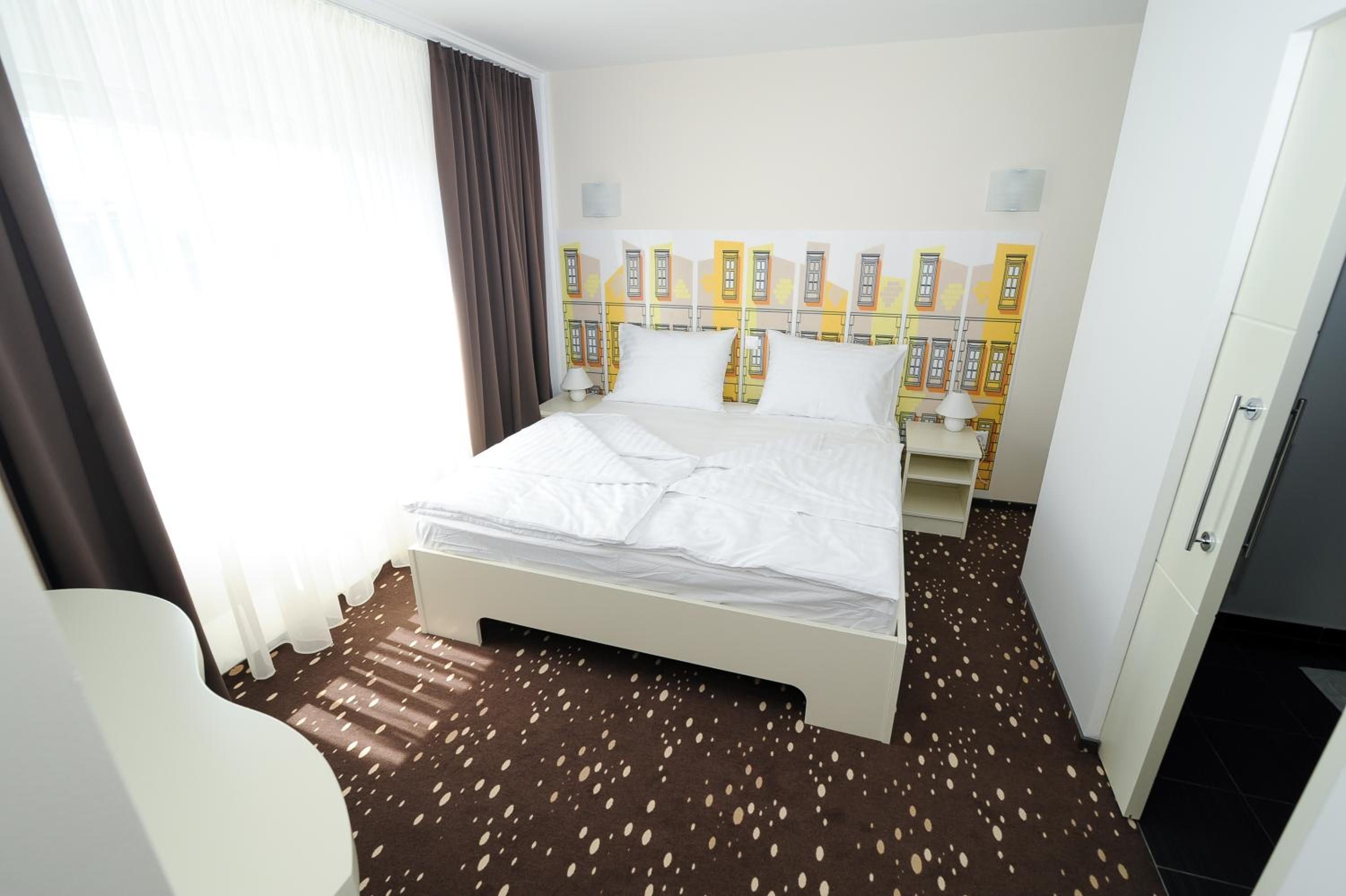 A hotel room with a bed, a chair, a wardrobe, and a wall with a cityscape mural.
