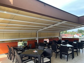 An outdoor patio area with tables and chairs, a covered roof, and a view of the surrounding area. - Hotel Gari A (Garesnica)