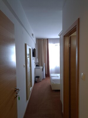 Quarto de hotel com cama, escrivaninha, cadeira, lixeira e janela com cortinas.