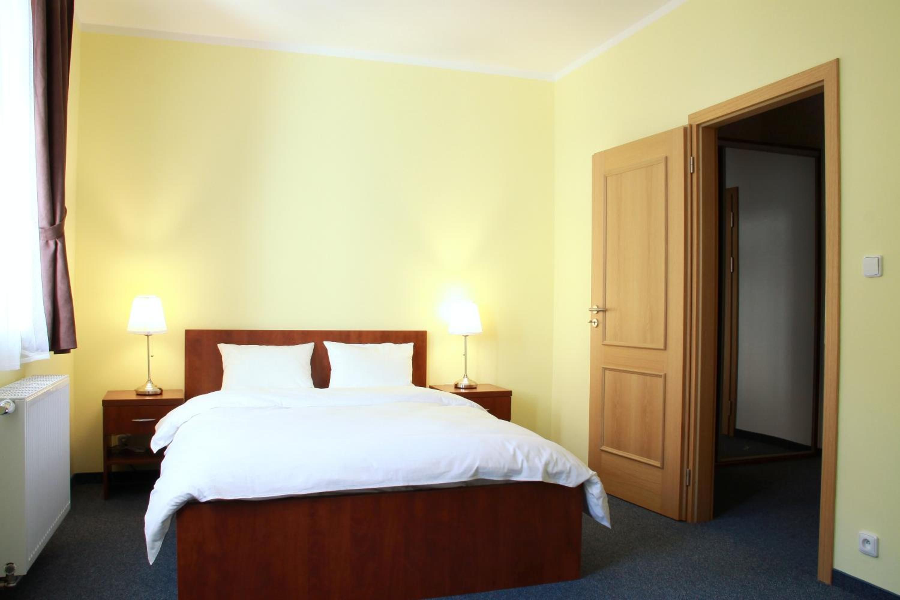 Ein Hotelzimmer mit einem Bett, zwei Nachttischen mit Lampen, einer Heizung und einer Tür zu einem anderen Raum.